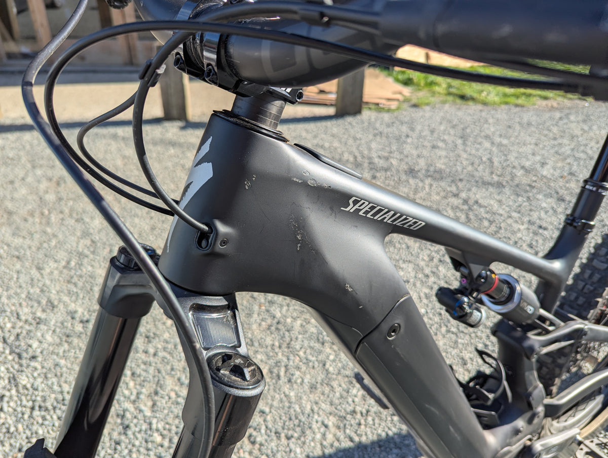 Close-up of a matte black Specialized full-suspension mountain bike frame, showing the brand name on the downtube and a Fox suspension fork, photographed outdoors on a gravel surface.