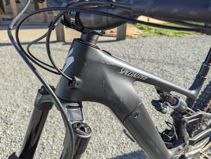 Close-up of a matte black Specialized full-suspension mountain bike frame, showing the brand name on the downtube and a Fox suspension fork, photographed outdoors on a gravel surface.