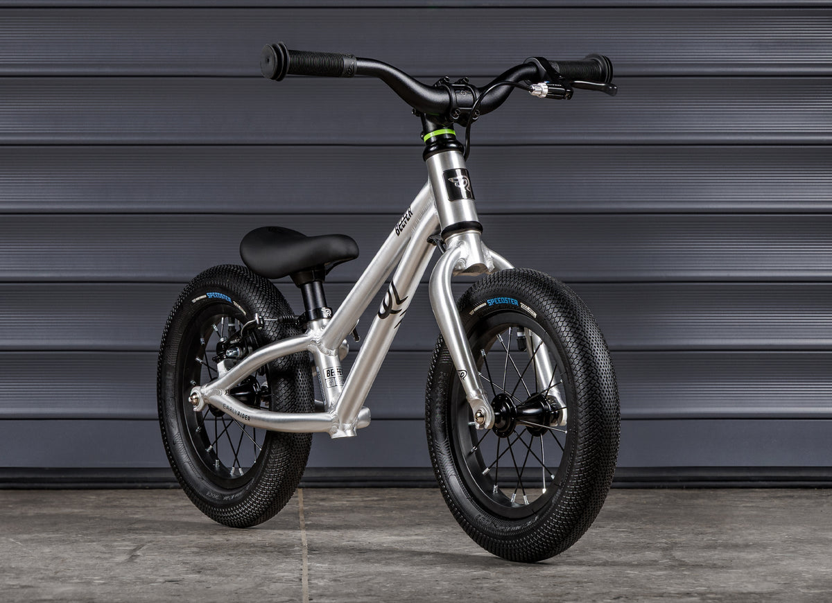 Early Rider Belter 12 Balance Bike in Brushed Aluminium - brushed balance bike against shutters, rear rim brake fitted