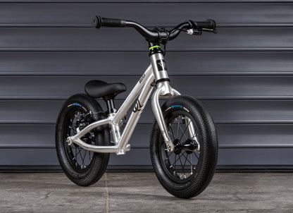 Early Rider Belter 12 Balance Bike in Brushed Aluminium - brushed balance bike against shutters, rear rim brake fitted