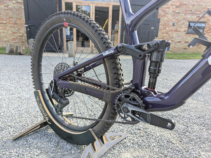 Close-up rear section of a purple and black full-suspension mountain bike on a stand, photographed outdoors on a gravel surface with a brick building in the background.