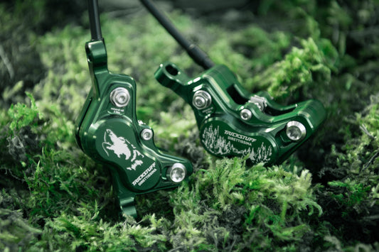 Two green Trickstuff Direttissima calipers with wolf and forest graphics resting on moss.